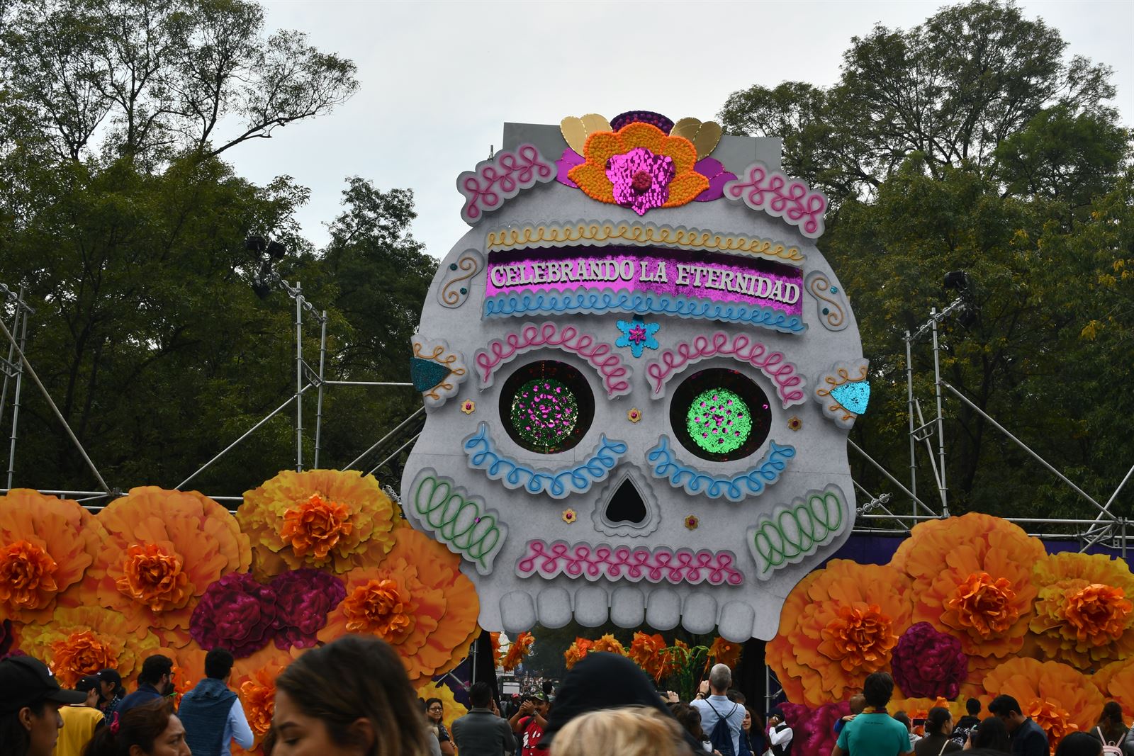 A giant decorated sugar skull float topped with paper marigolds reading Celebrando la Eternidad, above a crowd at a Dia de Muertos parade in Mexico